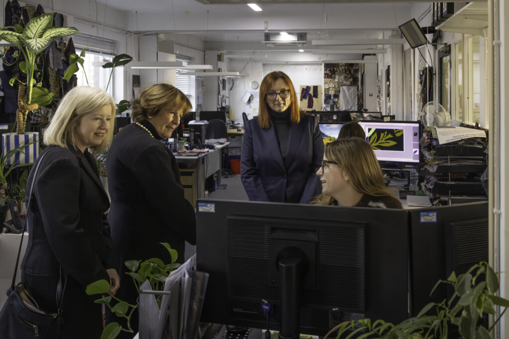 Three women speaking with designer and looking at her screen as they stand in a design studio