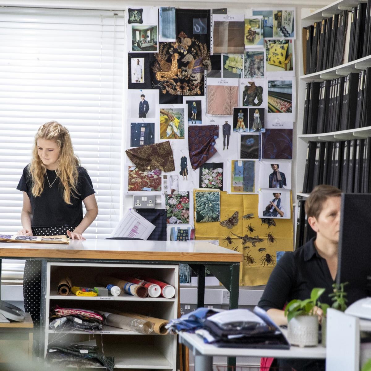 Two designers in the design studio. One at the far table looking at an archive book and one working at her desk. On the far wall is a moodboard pinboard of inspiration images.